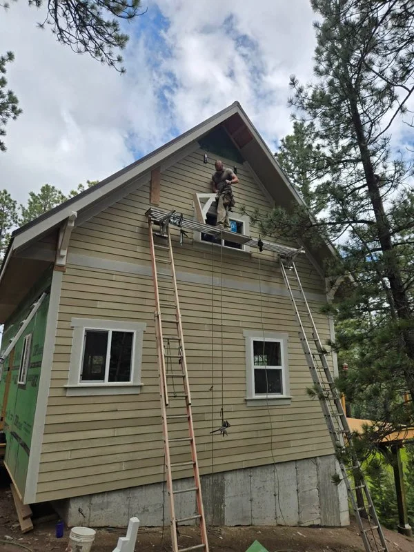 Two-story building with tan lap siding, white-trimmed windows, and a worker on a ladder-supported scaffold installing siding