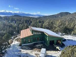 Mountain home with green siding, metal roof, and stone foundation surrounded by forest
