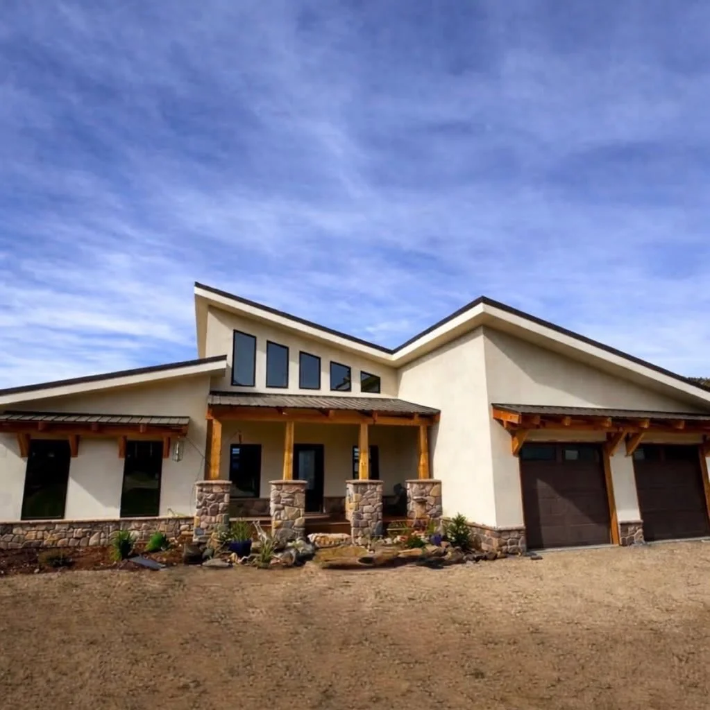 Stucco home with stacked stone columns, exposed timber beams, dark metal roofing, and black-framed windows