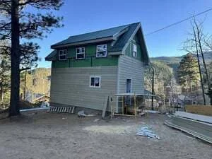 Elevated house with green and gray siding, pitched roof, concrete pilings, and gravel foundation