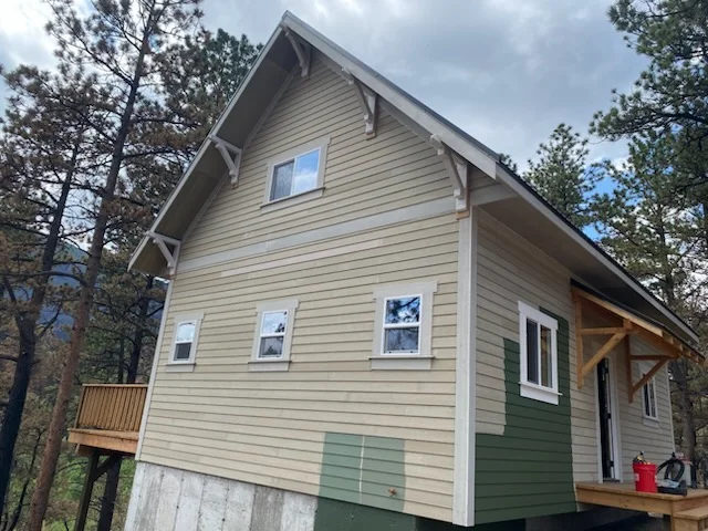 Two-story cabin with cream lap siding, sage green panel accents, timber brackets, and a covered timber entry porch